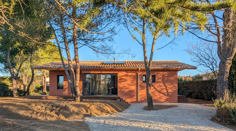 Maison de plain-pied avec façade ocre, toit en tuiles et panneaux solaires. Une terrasse en bois est aménagée devant la maison, avec quelques pots de fleurs. Le bâtiment est entouré d'arbres, créant une ambiance naturelle et chaleureuse