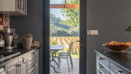 Cuisine moderne avec des meubles gris et un plan de travail en marbre, offrant une vue directe sur une terrasse extérieure en bois et un vignoble à travers une porte vitrée. La lumière naturelle éclaire l'espace, créant une ambiance chaleureuse. La table et les chaises sur la terrasse offrent un cadre idéal pour profiter de l'extérieur, entouré par les rangées de vignes. Le contraste entre les teintes sombres de l'intérieur et la verdure extérieure renforce l'harmonie entre l'espace intérieur et extérieur.