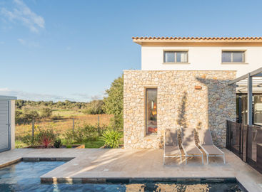 Maison moderne avec une façade en pierre naturelle, située au bord d'une piscine. La terrasse en pierre offre un espace de détente équipé de chaises longues. La fenêtre étroite laisse entrer la lumière naturelle tout en préservant l'intimité de l'intérieur. Le paysage environnant, composé de champs ouverts et de végétation, accentue l'ambiance sereine et naturelle de la propriété. Le toit en tuiles et les finitions en pierre soulignent l'harmonie entre modernité et matériaux traditionnels.