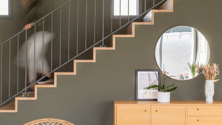 Intérieur lumineux avec un salon au design contemporain, comprenant un escalier suspendu avec des marches en bois et un garde-corps métallique. Le mur d'accent est peint en gris foncé, contrastant avec le mobilier minimaliste en bois clair, notamment un buffet et une table basse. Le plafond en bois lamellé apporte une touche chaleureuse à l'espace, tandis que le sol en carrelage blanc renforce la sensation de luminosité et d'espace. Une chaise en rotin et des touches décoratives comme un miroir rond et des étagères colorées ajoutent des éléments de texture et de contraste à cet intérieur moderne.
