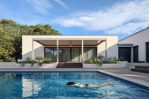 Maison contemporaine avec toiture plate, large auvent formant une terrasse couverte. La façade est équipée de grandes baies vitrées à double vitrage qui s'ouvrent sur une terrasse en bois. Une piscine à débordement se situe en premier plan, reflétant l'architecture épurée de la maison. Le contraste entre les lignes rectilignes de la construction et la végétation environnante met en valeur l'intégration harmonieuse du bâtiment dans son environnement naturel.