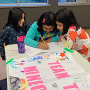 Three young girls sitting at a table with a sign on it, happily captivated in their drawings.