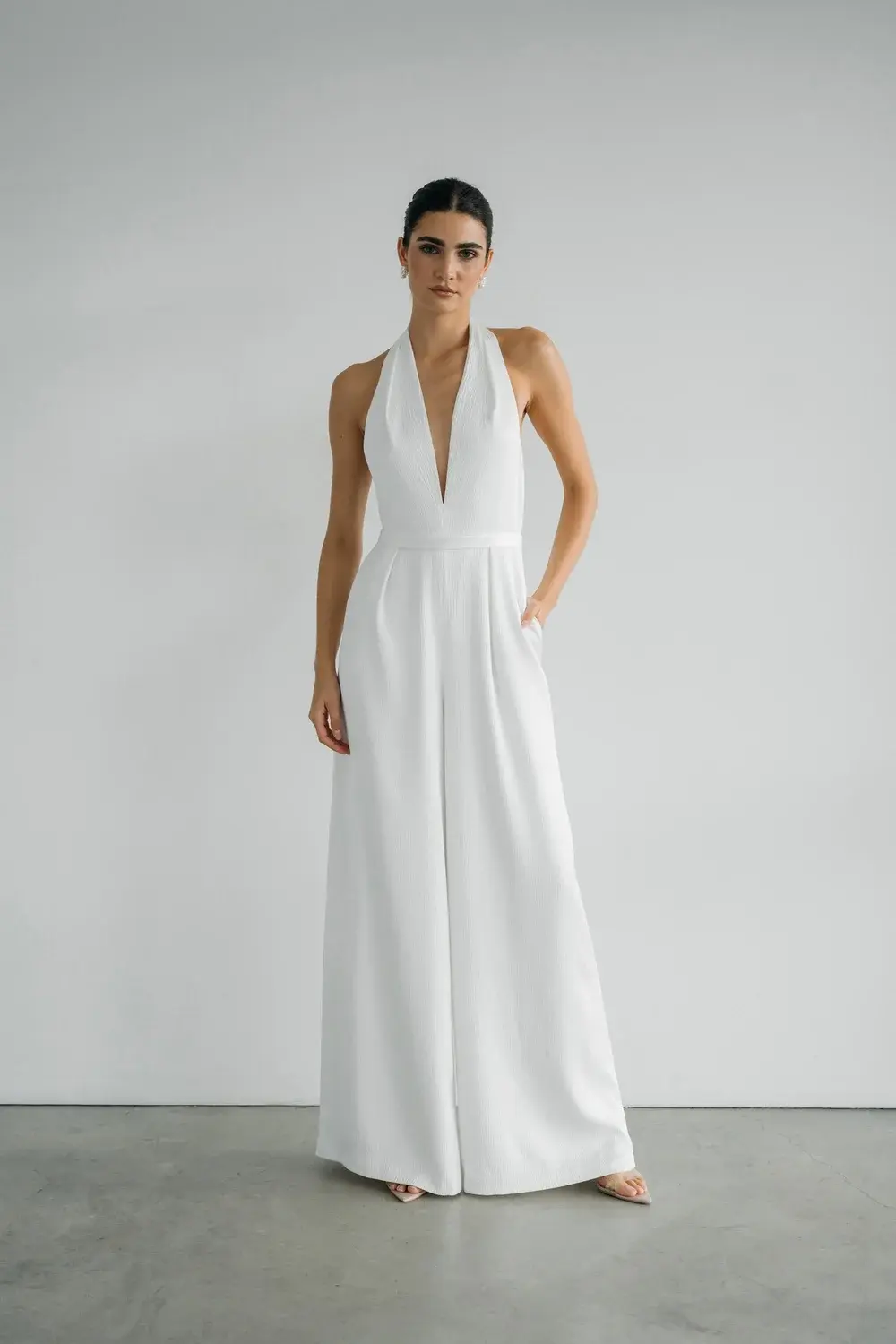 Woman wearing white halter jumpsuit with pockets, standing against a white backdrop.