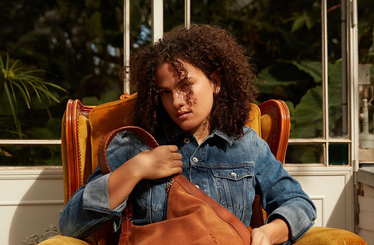 Jeune femme assise dans un fauteuil de style western avec une veste en jean et un sac en cuir camel Paul Marius.