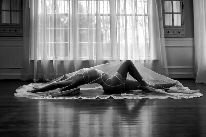 Black and white boudoir portrait of a woman in lace lingerie lying beneath a sheer bridal veil, backlit by large window light in an elegant interior setting.
