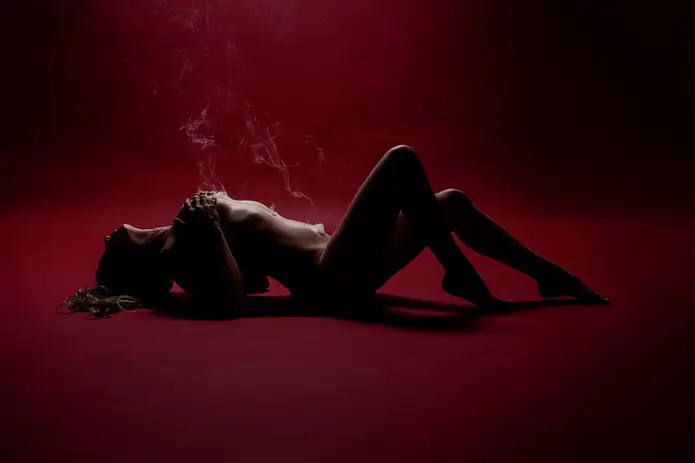 Silhouette boudoir portrait of a woman reclining against a red background with dramatic lighting and rising smoke creating a bold, artistic effect.