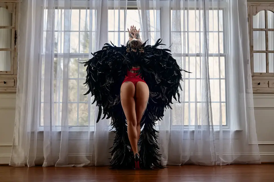 Boudoir portrait of a woman wearing red lingerie and large black feathered wings, standing in front of a bright window with sheer curtains.