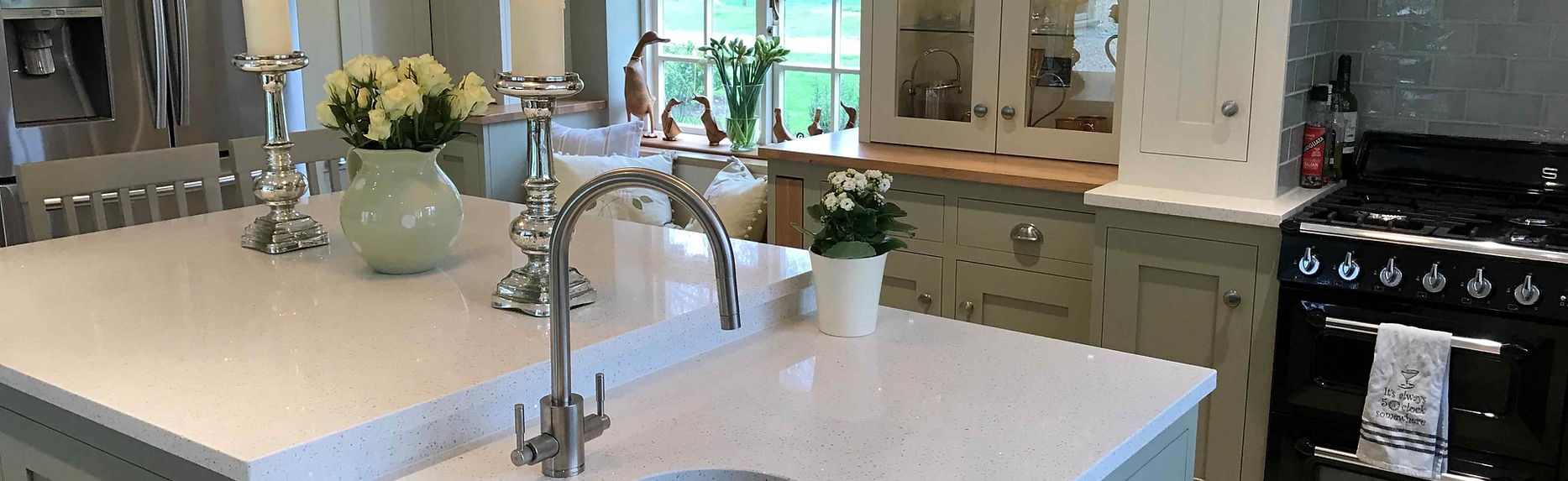 Elegant sage green shaker kitchen featuring a central island with a small prep sink, quartz worktops, and chrome accessories in a light-filled country home.