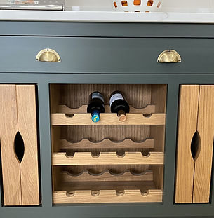 Natural oak wine rack integrated into a green-painted base cabinet with brass cup handles, designed for horizontal bottle storage in a classic kitchen.
