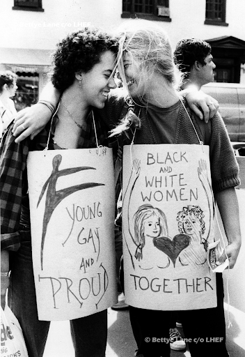 Alt Text: Two women with signs hung around their neck hold a single arm embrace and touch foreheads smiling.