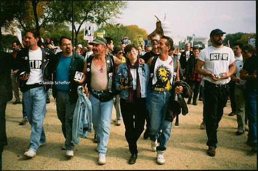 Five Act Up Activists with their arms around each other, walking in a straight line and carrying ashes in boxes, another activist wearing an act up shirt, all marching together.