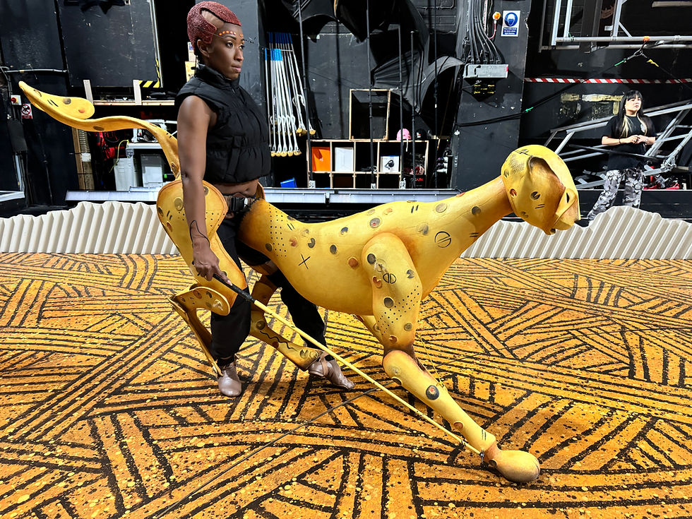 Person manipulating a cheetah puppet in a patterned room. Puppeteer looks focused. Background has equipment and another person standing.