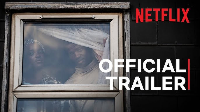 Two people look out a window, one pulling back a curtain. The words "Netflix Official Trailer" are displayed. The mood is tense.