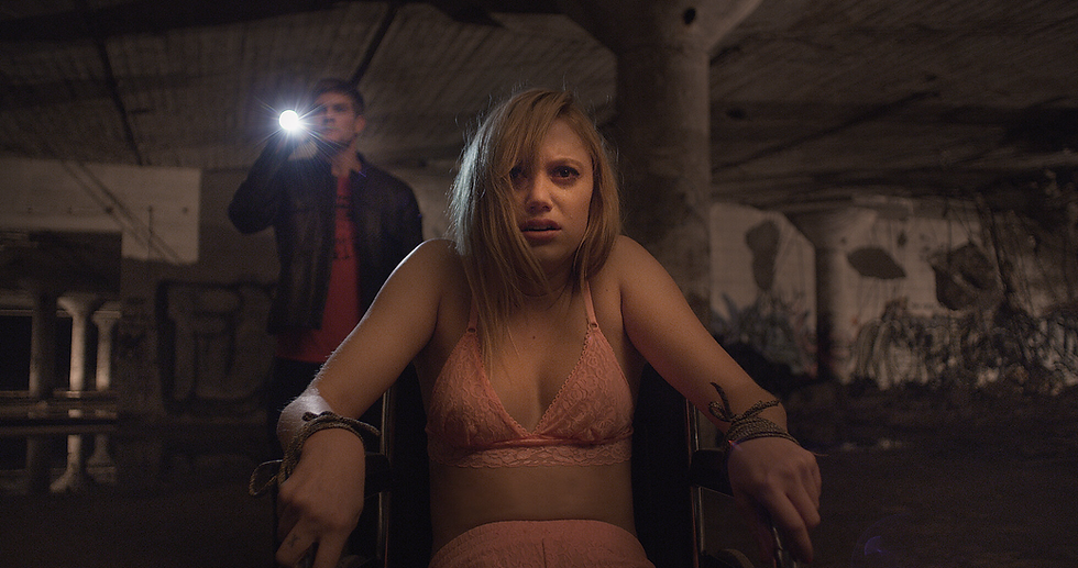 Woman in distress, tied to a chair in a dim, abandoned space. Man in the background holds a flashlight. Mood is tense and fearful.