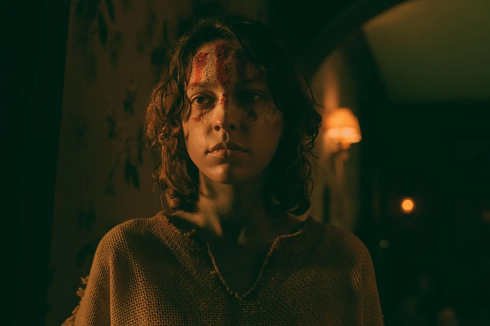 A person with a distressed expression stands in dim lighting, face marked with red paint, wearing a textured brown shirt. Mood is tense.