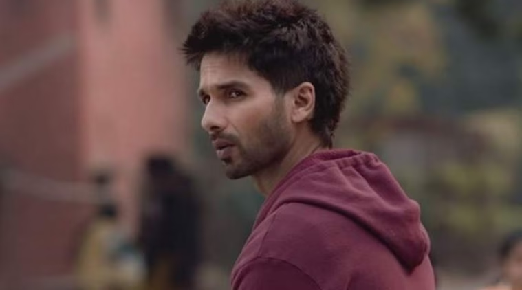 Man in a maroon hoodie looks thoughtfully over his shoulder. Blurred outdoor background with subtle greenery and a brick wall.