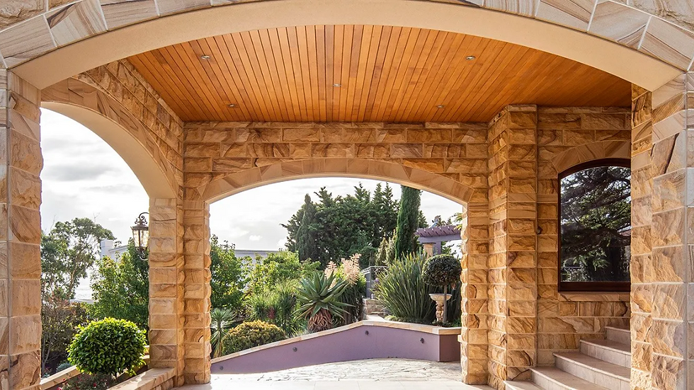 Stone archway with a wooden ceiling overlooks a lush garden. Pillars and walls are made of textured sandstone. Bright, peaceful ambiance.