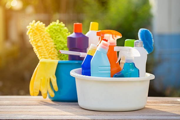 Cleaning supplies in colorful bottles and a mop in buckets on a wooden table, outdoors. Gloves and brush visible, in warm sunlight.
