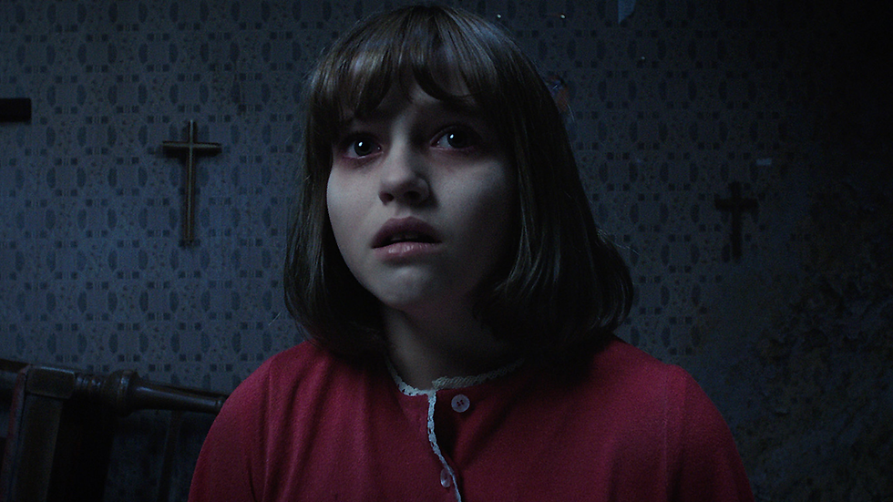 Girl in red shirt looks upward with a fearful expression. Dimly lit room with crosses on patterned wallpaper conveys a tense atmosphere.