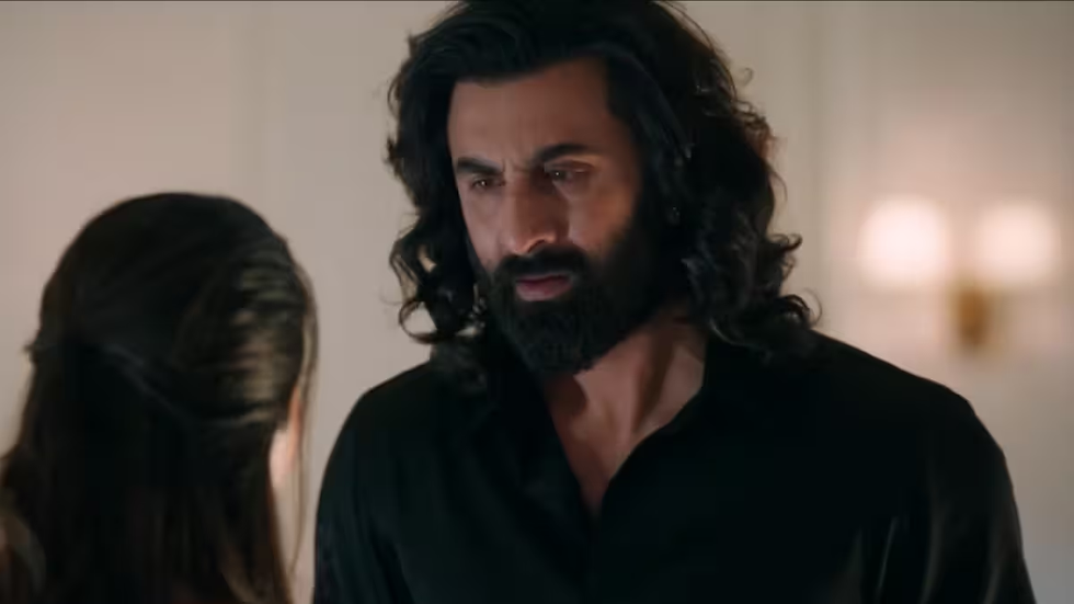 Man in black shirt looks serious in a softly lit room, facing a woman with her back to the camera. Warm lighting and neutral background.