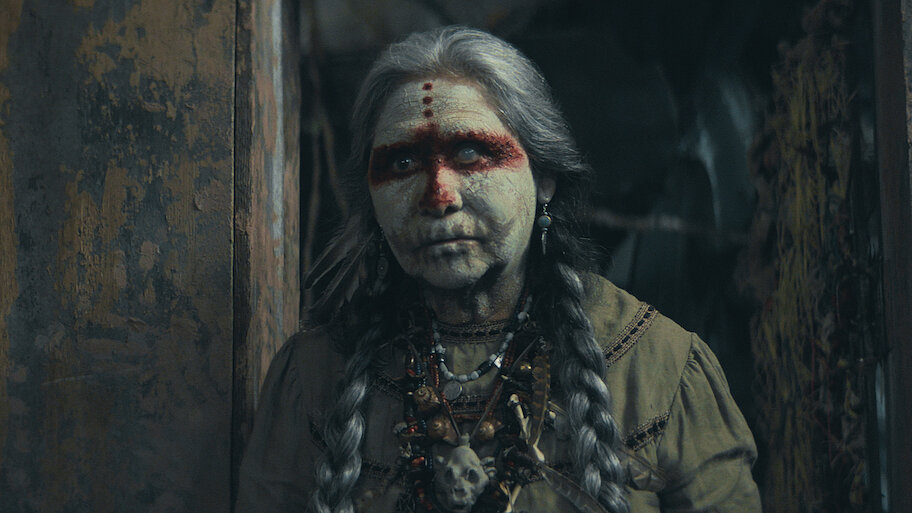 Elderly woman with braided hair and painted face, wearing a beaded necklace, stands in dimly lit rustic setting, conveying a solemn mood.