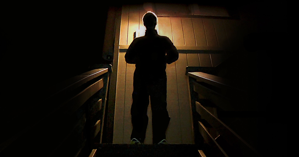 Silhouetted figure stands at the top of dimly-lit stairs facing a wooden door. Warm light creates a mysterious, suspenseful mood.