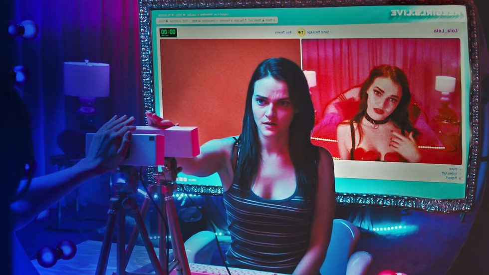 Woman in striped tank top operates a camera in a dim room. Her reflection in the screen shows her in red with a different expression.