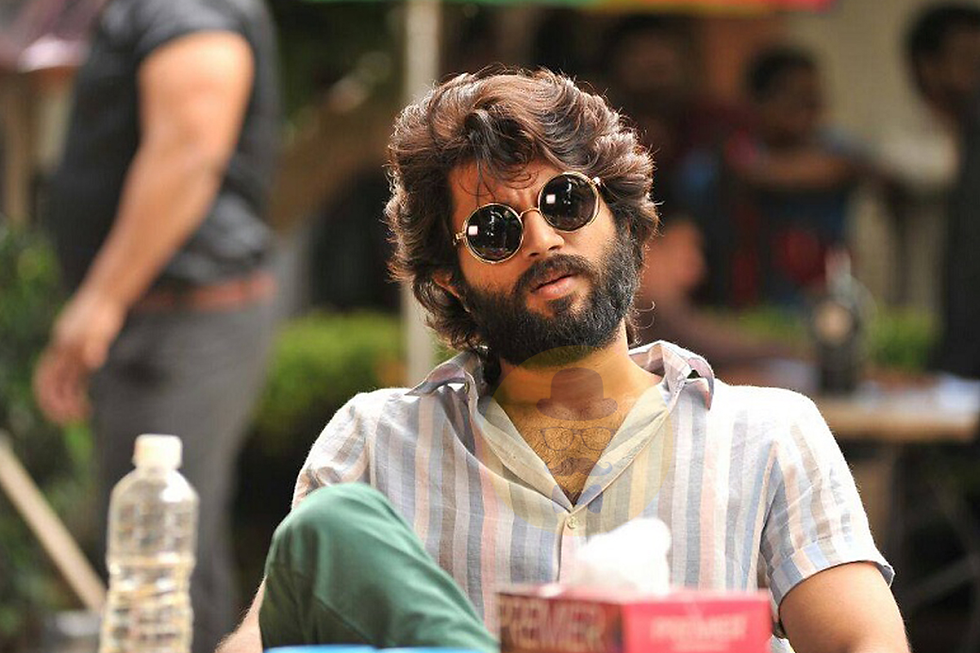 Bearded man in sunglasses and striped shirt sits casually outdoors. Water bottle and red box on table. Relaxed, sunny ambiance.