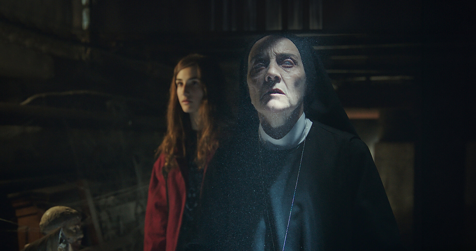 Nun in black habit with serious expression stands beside a young woman in a red jacket. Dim, mysterious setting with dramatic lighting.