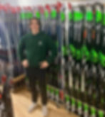 Un employé du Domaine Saint-Bernard sourit à la caméra devant un mur de skis de fond à louer