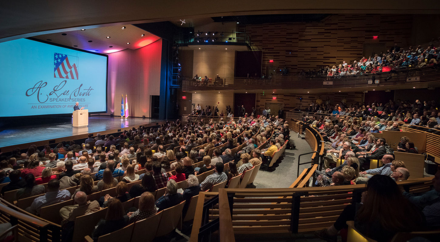An interior view of the Mainstage Theatre at the Bicknell Family Center for the Arts. Looking from House Left towards the stage. The auditorium is full, and a podium with a speaker is at Center Stage.