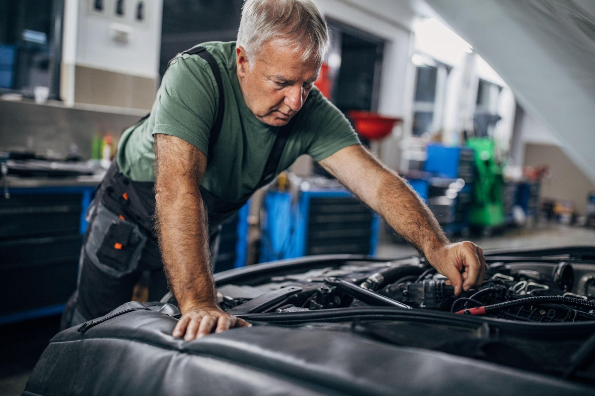 Older mechanic in green shirt leans over an open car hood in a garage, carefully inspecting the engine with a focused look.
