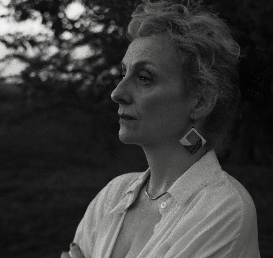 "Plano médio em preto e branco da personagem Myriam no filme 'A Mensageira'. Ela aparece de perfil, com expressão pensativa, usando brincos geométricos e camisa clara, em um cenário externo sombreado."