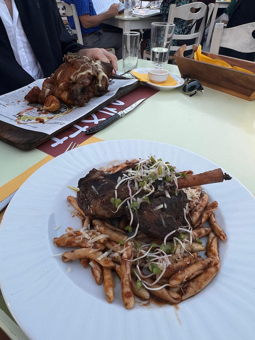 Outdoor restaurant table with plated lamb shank, greek pasta and sprouts; another roast sits behind amid diners and glasses.