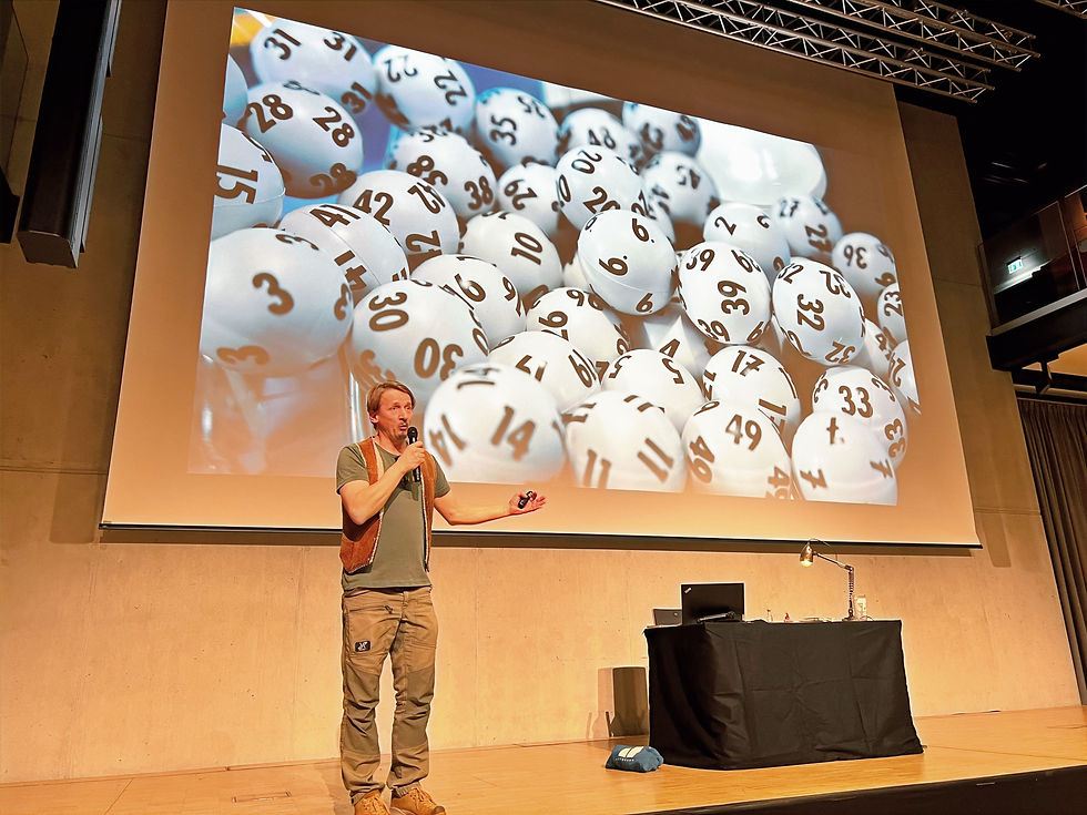 Oliver Lück erzählt auf der Bühne der PS.Halle im PS.SPEICHER mit Mikrofon eine Geschichte und zeigt dabei humorvolle Reisebeobachtungen. Im Hintergrund wird auf der Leinwand eine Präsentation mit Lotto-Bällen gezeigt.