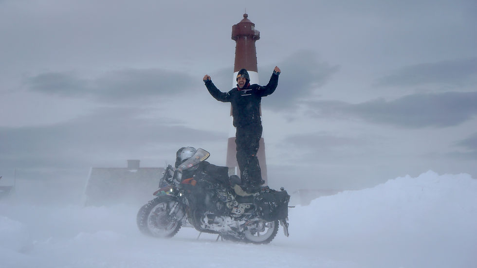 Bruno Pillitteri steht jubelnd auf seinem verschneiten Motorrad vor dem Slettnes-Leuchtturm, während starker Wind Schnee durch die winterliche Landschaft treibt.