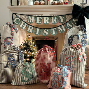 A collection of striped Christmas sacks with a Liberty printed floral initial on each one and Merry Christmas bunting hung over a fireplace