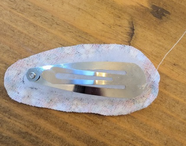 Metal hair clip with a white fabric backing, placed on a wooden surface. A thread extends from the right, suggesting handmade crafting.