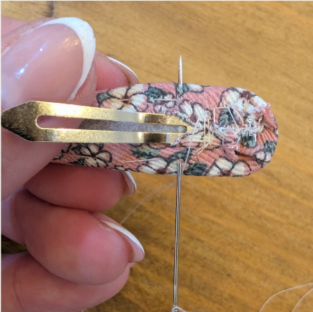 Hand sewing a floral fabric onto a hair clip with a needle and thread. Pink nails, wooden surface, intricate stitching in progress.