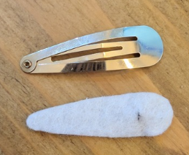 A metallic hair clip and a white felt piece lie on a wooden surface. The clip is shiny, reflecting light, creating a simple and clean look.