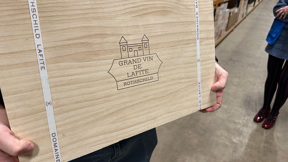 Person holds a wooden crate labeled "Grand Vin de Lafite Rothschild" in a warehouse filled with stacked boxes. Beige and earthy tones.