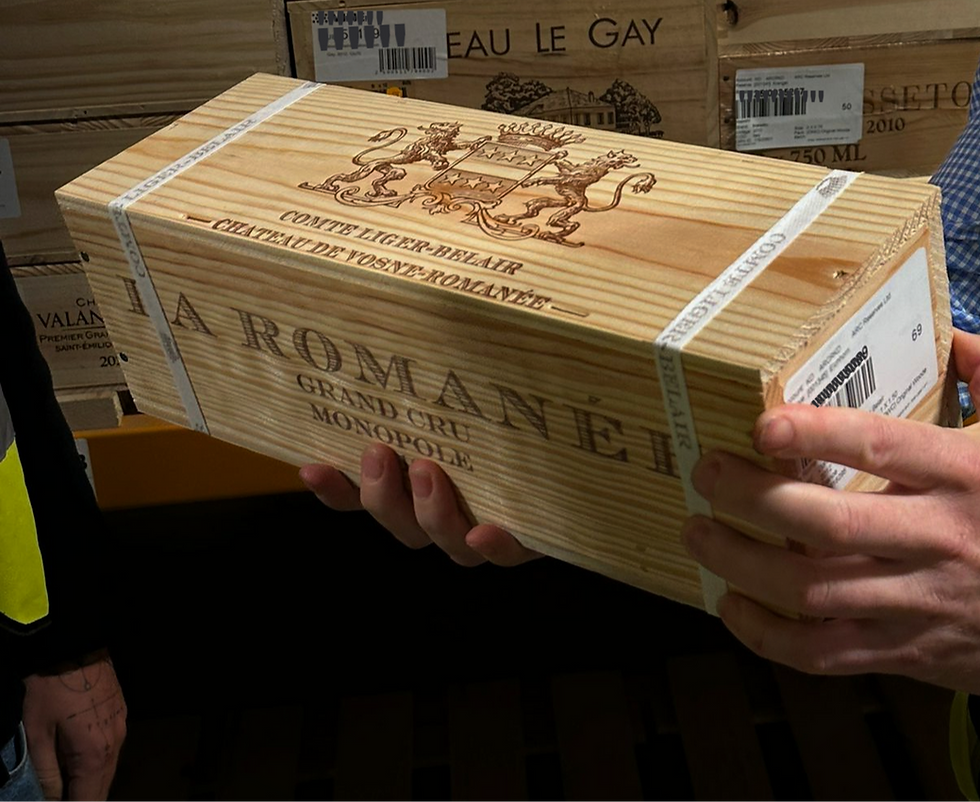 Hands holding a wooden wine crate labeled "LA ROMANÉE GRAND CRU MONOPOLE," with a lion crest. Background shows stacked wooden wine boxes.