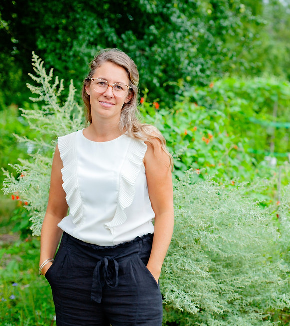A woman wearing a white shirt, glasses, and grey pants. She is standing in a field of tall grass and flowers.
