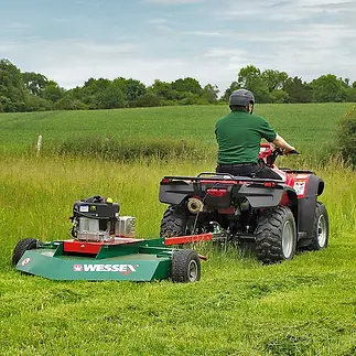 A man on a red quad bike towing a green Wessex rotary mower to cut long grass in a large open field