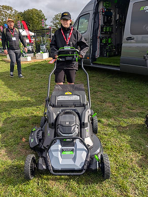 A person in a black jacket standing behind a grey EGO Power+ cordless mower at an outdoor trade show