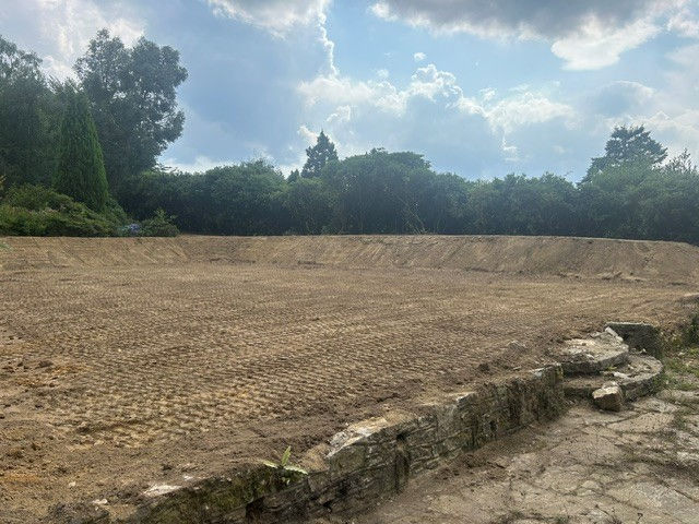 Freshly graded dirt field bordered by stone wall and trees