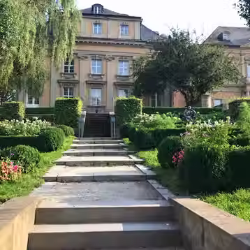 Stone steps lead between clipped hedges and flower beds toward a yellow historic façade in Bayreuth.