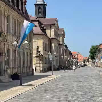Sunny cobbled street beside historic sandstone buildings and a church tower in Bayreuth.