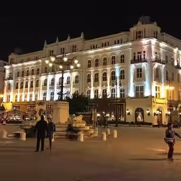 Illuminated Gerbeaud House behind a stone lion fountain and pedestrians in Vörösmarty Square, Budapest.