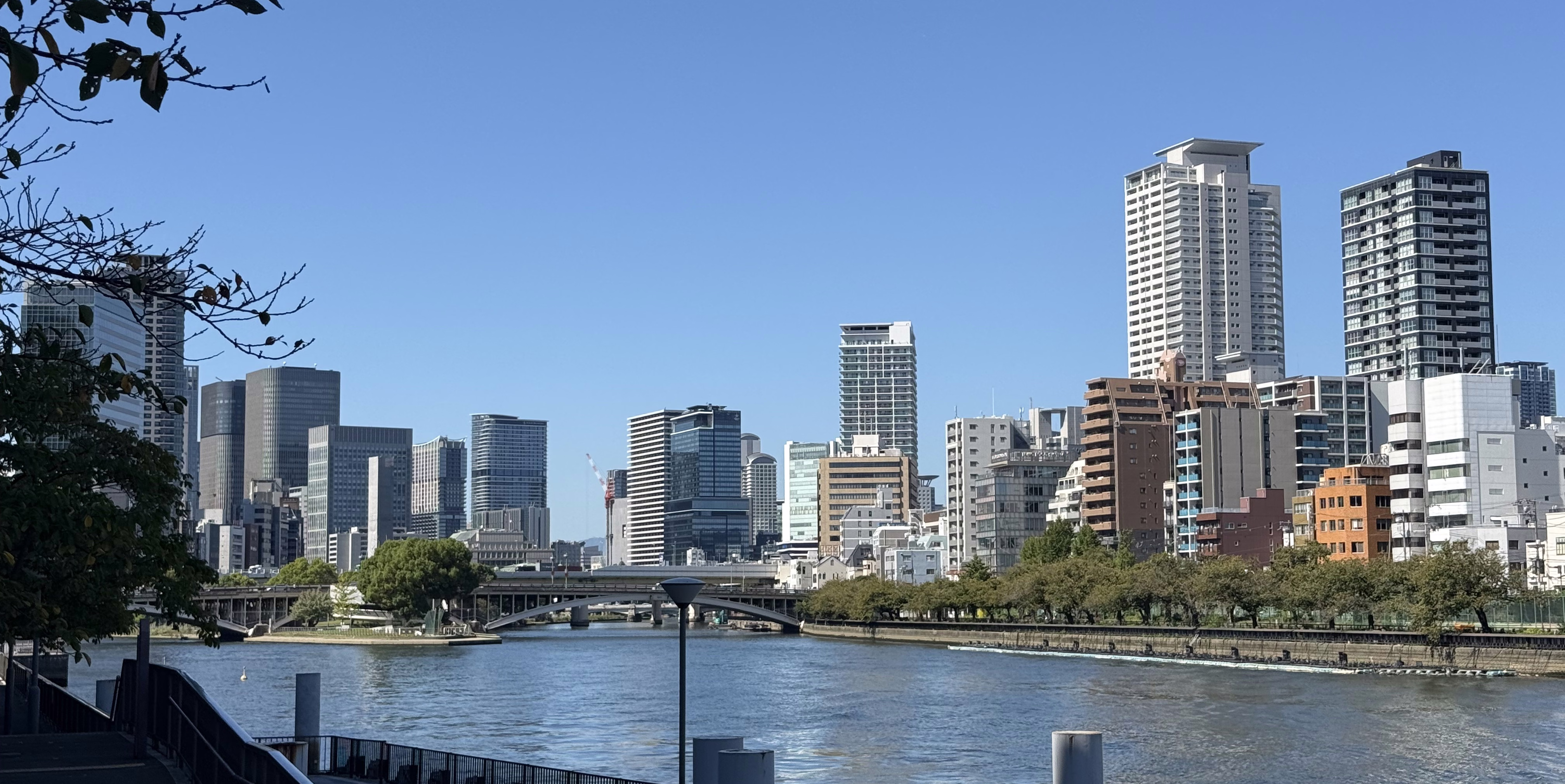 Osaka riverfront skyline with bridges, waterside trees and high-rise buildings under a clear blue sky.